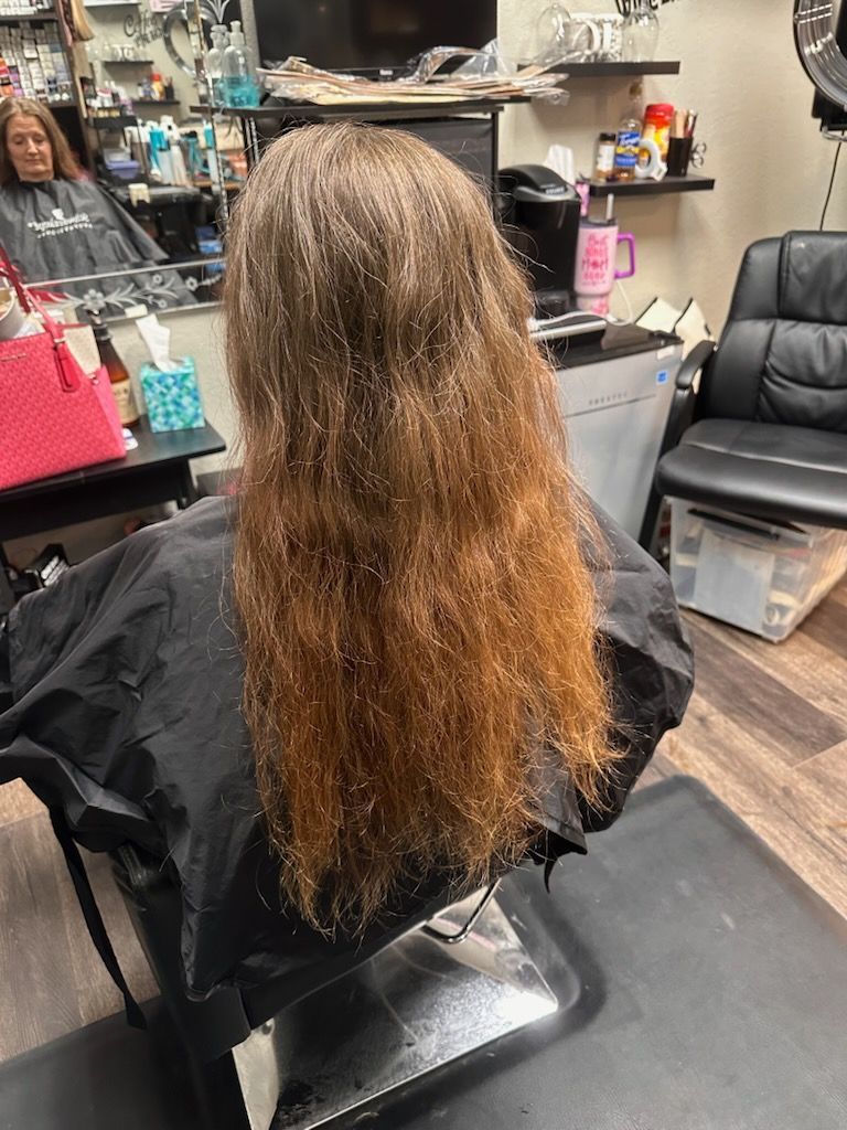 Person seated in a salon chair with long, wavy brown hair, facing a mirror and styling tools.