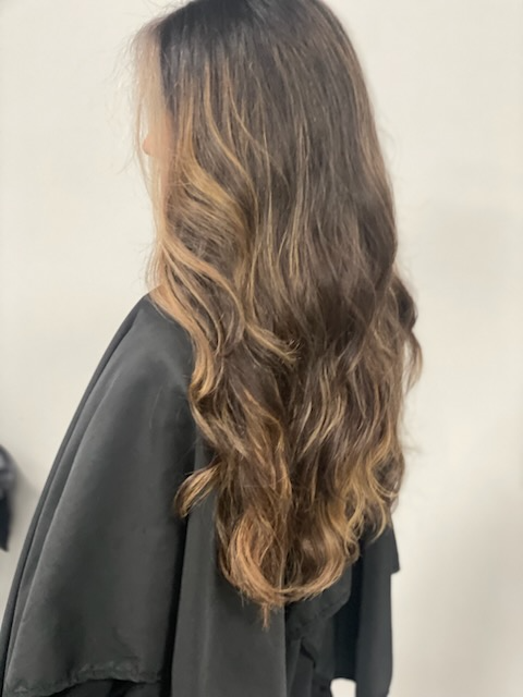 Woman with long wavy brown hair wearing a black cape, shown from behind against a light wall