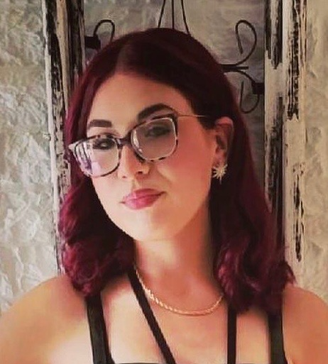 Portrait of a person with burgundy hair, glasses, and a black top against a textured wall.