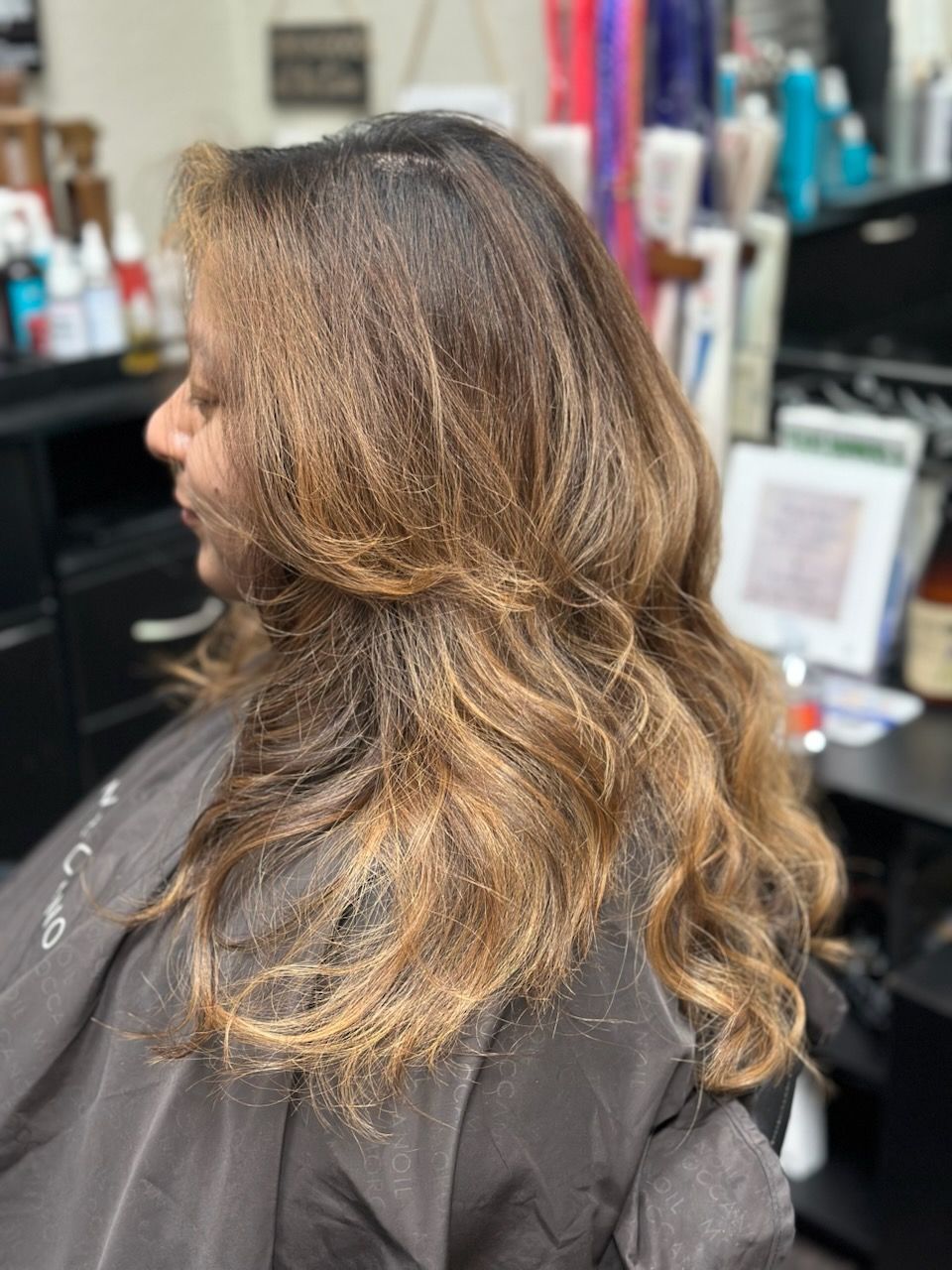 Side view of a person with wavy brown hair in a salon chair, showing layered highlights.