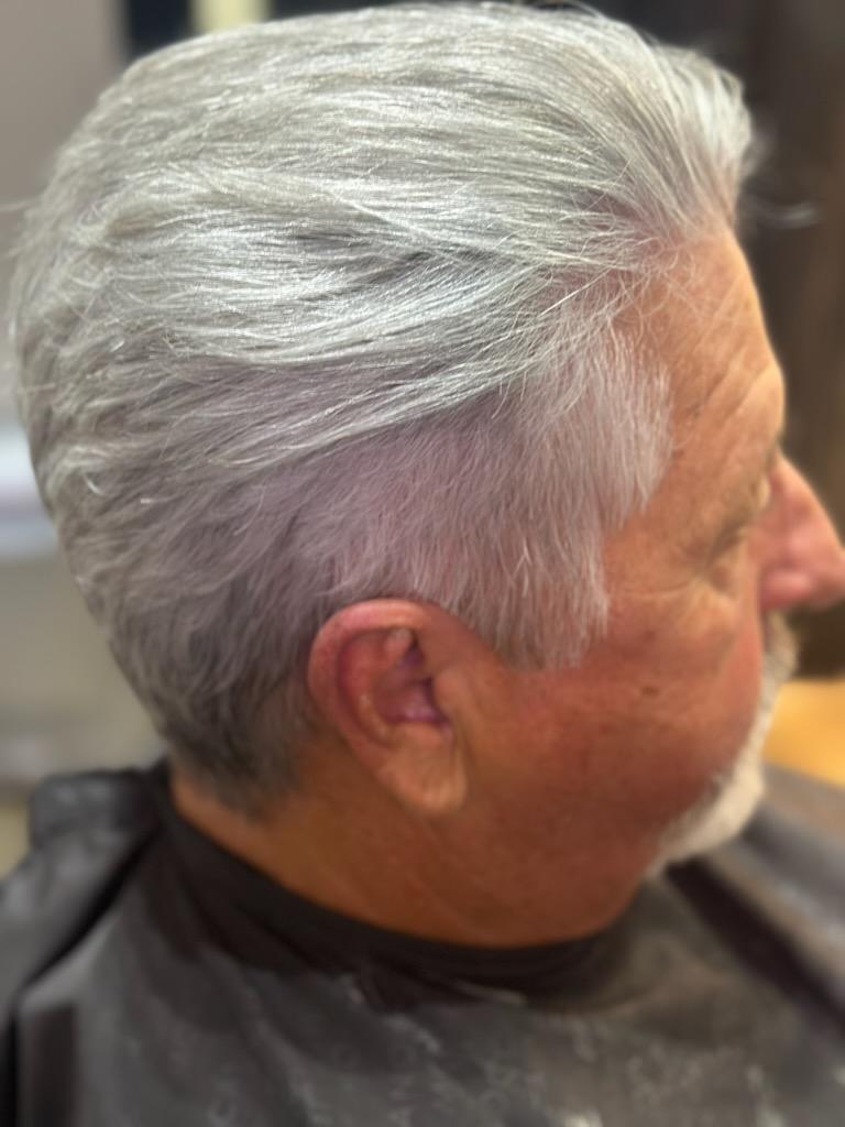 Profile view of a man with neatly combed silver hair seated in a salon chair, wearing a black cape