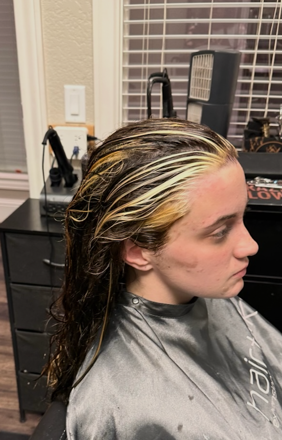 Person in a salon cape with wet, blonde-highlighted hair, seated in a chair.