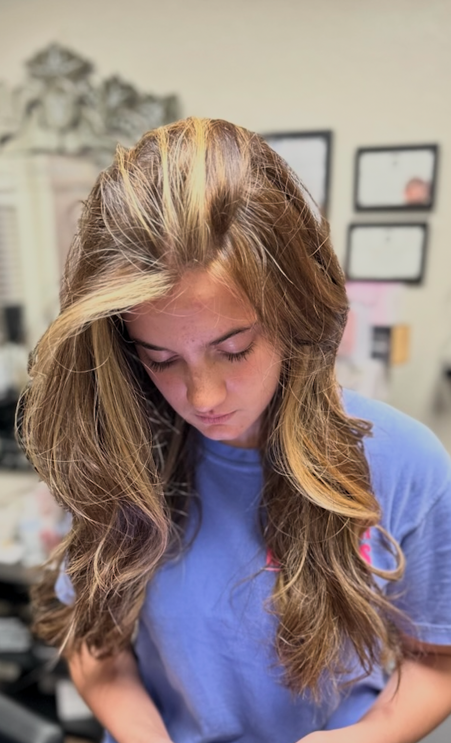 Person with long hair looking down in a blue shirt indoors, with framed wall art in the background
