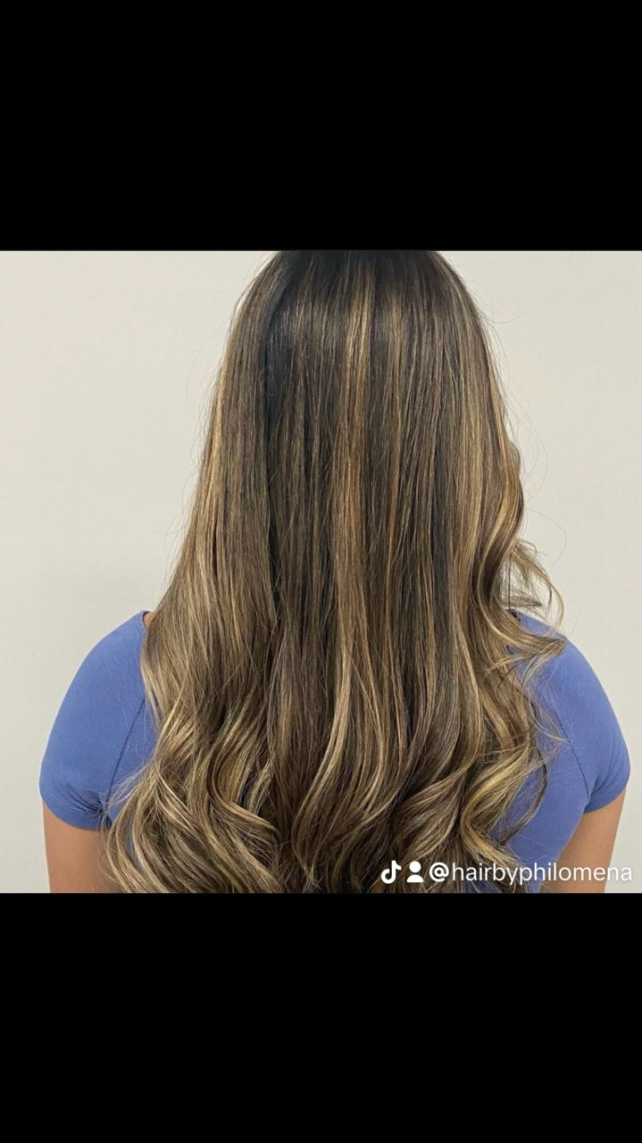 Back view of long wavy brown-highlighted hair over a blue shirt against a plain wall
