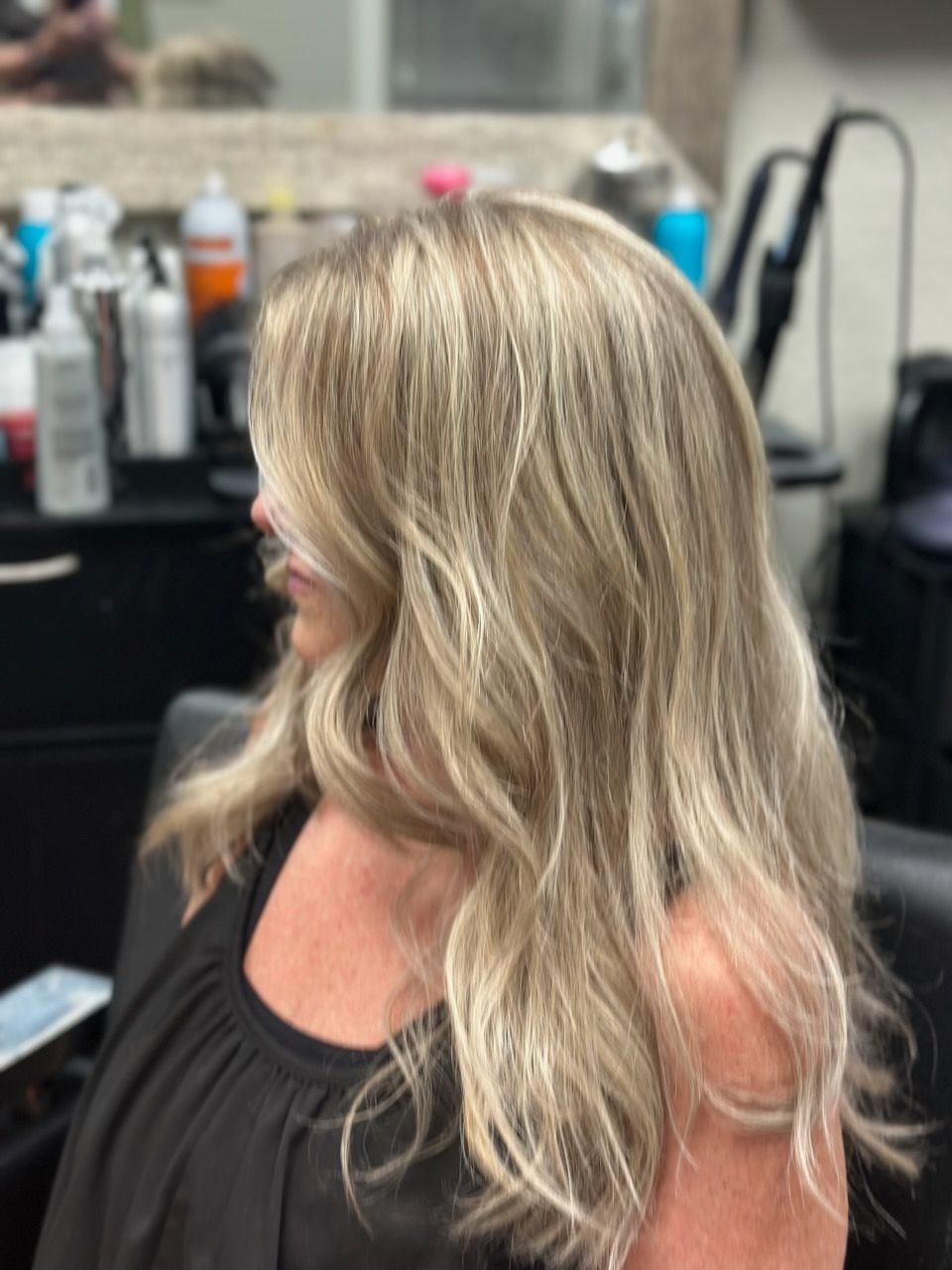 Woman with long blonde hair and soft waves, seated in a salon chair wearing a black top.