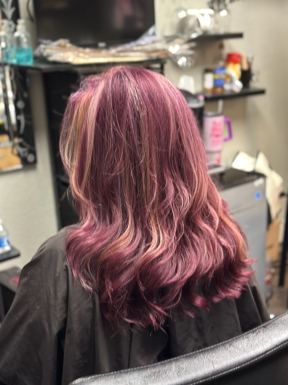 Salon client with shoulder-length wavy hair dyed burgundy and pink highlights, seen from behind.