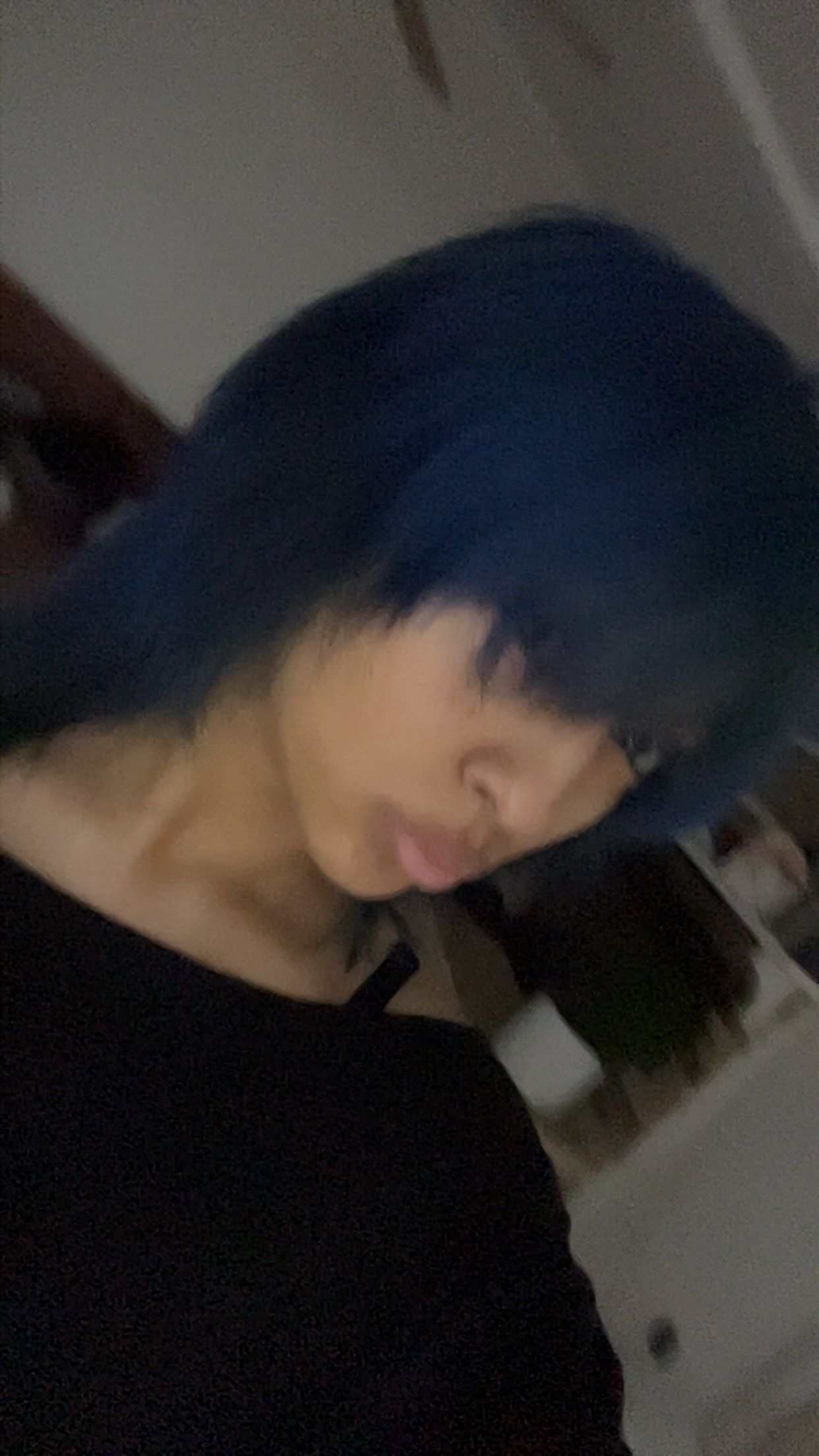 Person with short blue hair and black top looking down in a dim indoor setting