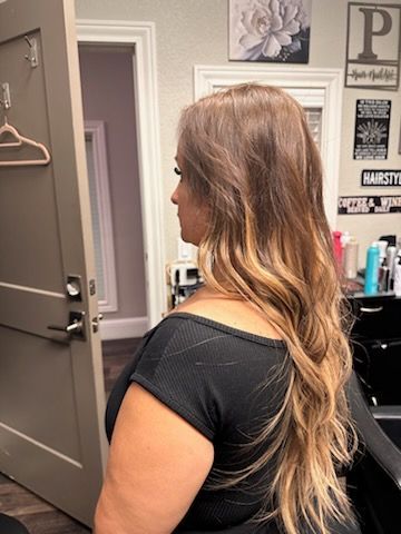 Woman with long wavy blonde hair in a salon chair, shown from the side in a black top.