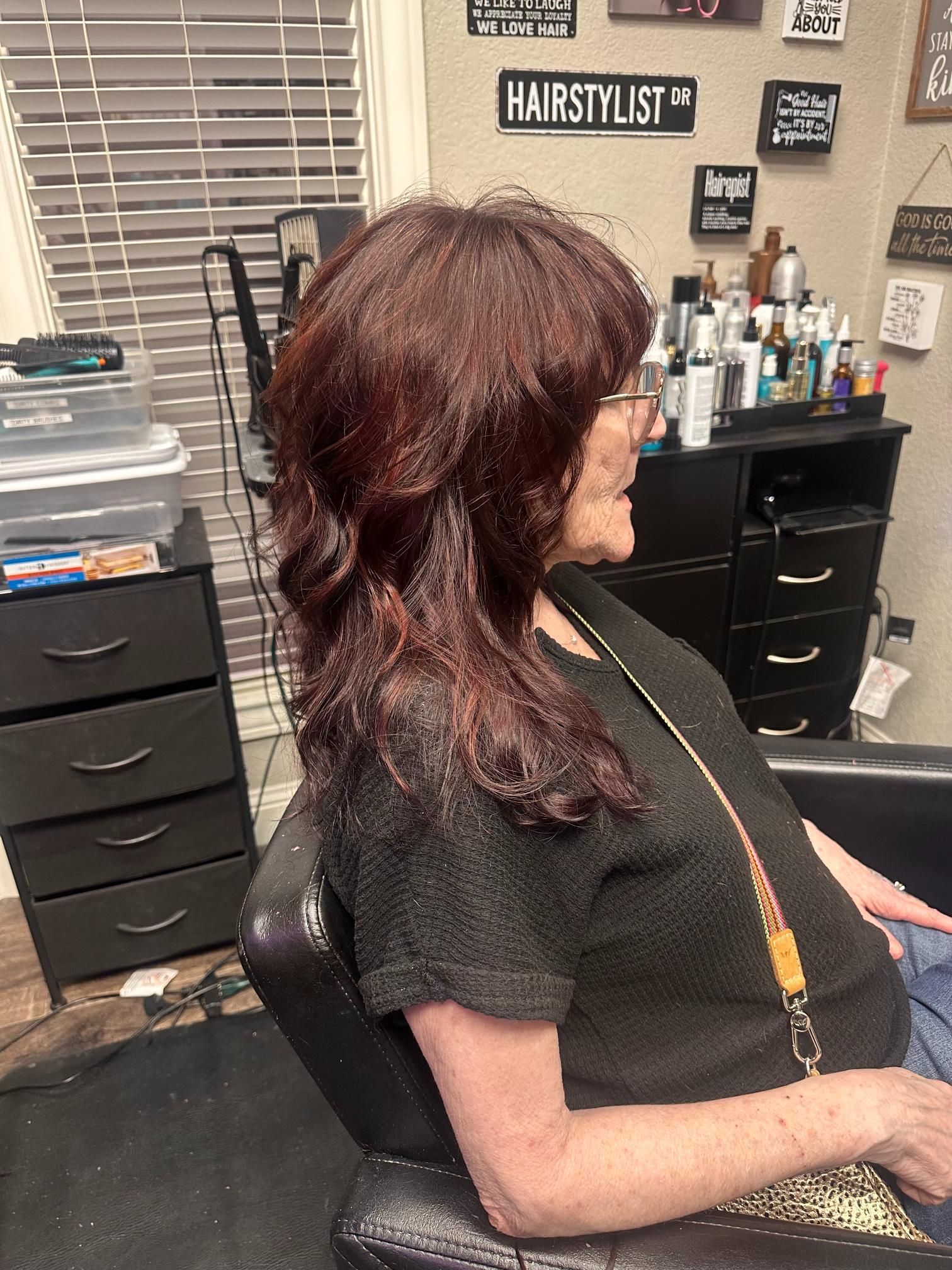 Woman with burgundy hair sitting in a salon chair, facing right, in a hairstyling studio.