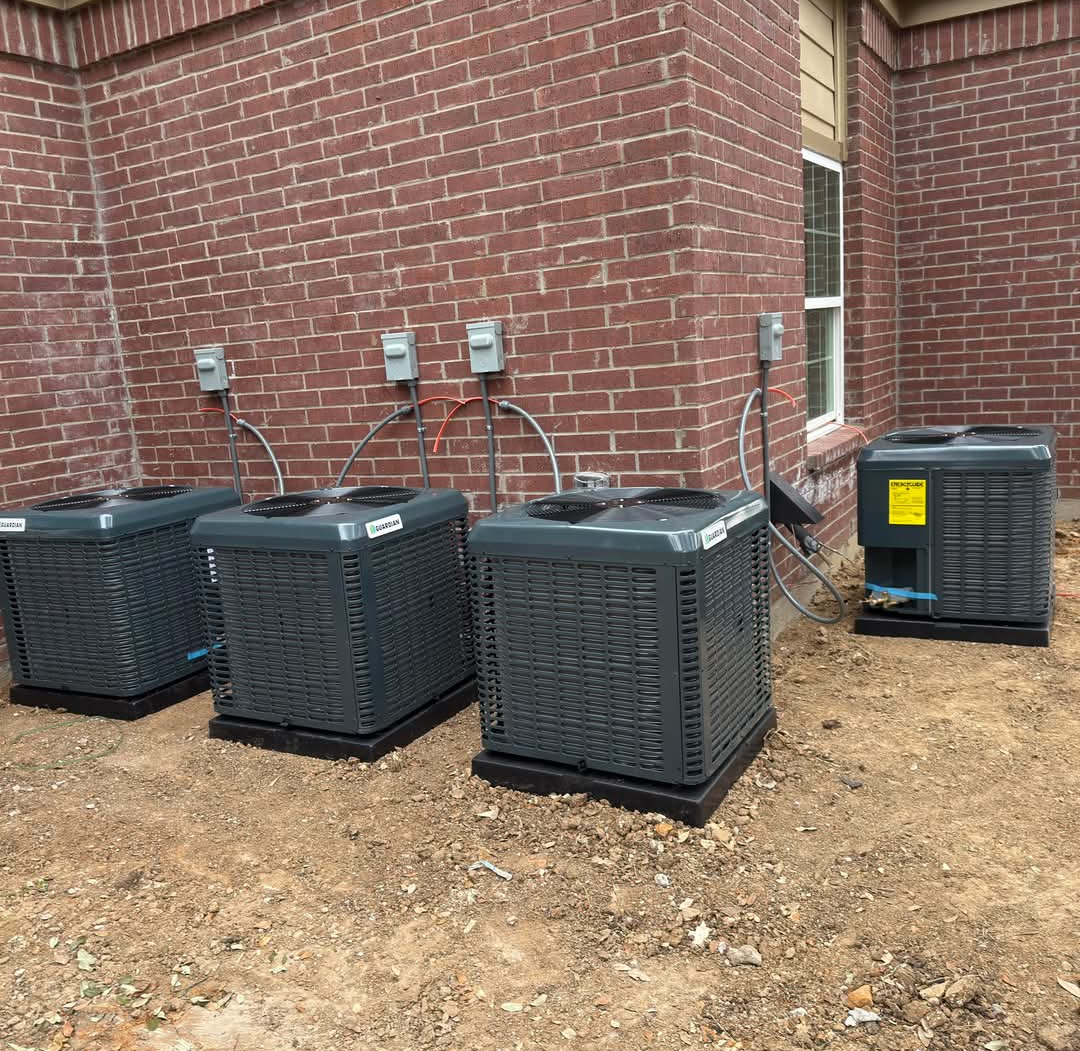 Four gray air conditioning units against a brick wall, connected to electrical boxes.