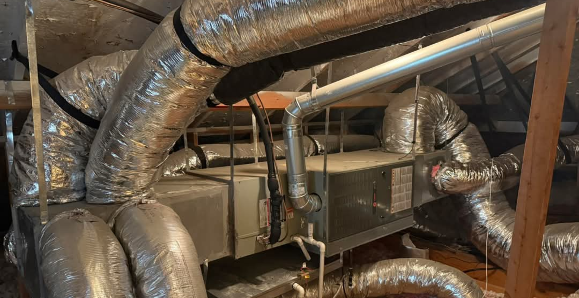 HVAC system in an attic with silver ductwork, gray unit, and wooden beams.