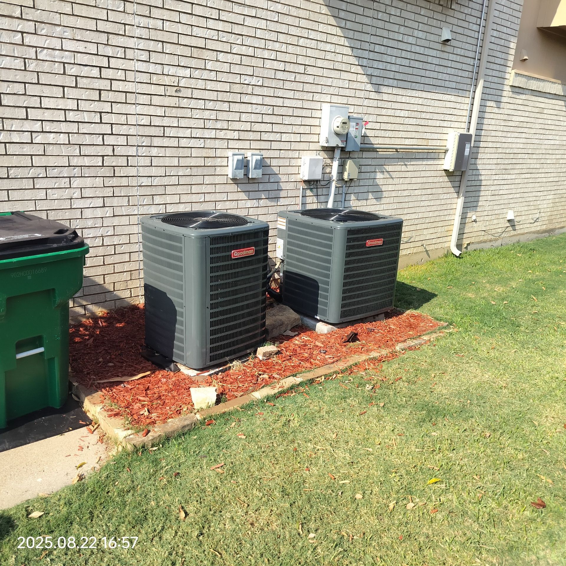 Two air conditioning units outside a brick building, near a trash can. Red mulch in the flower bed.