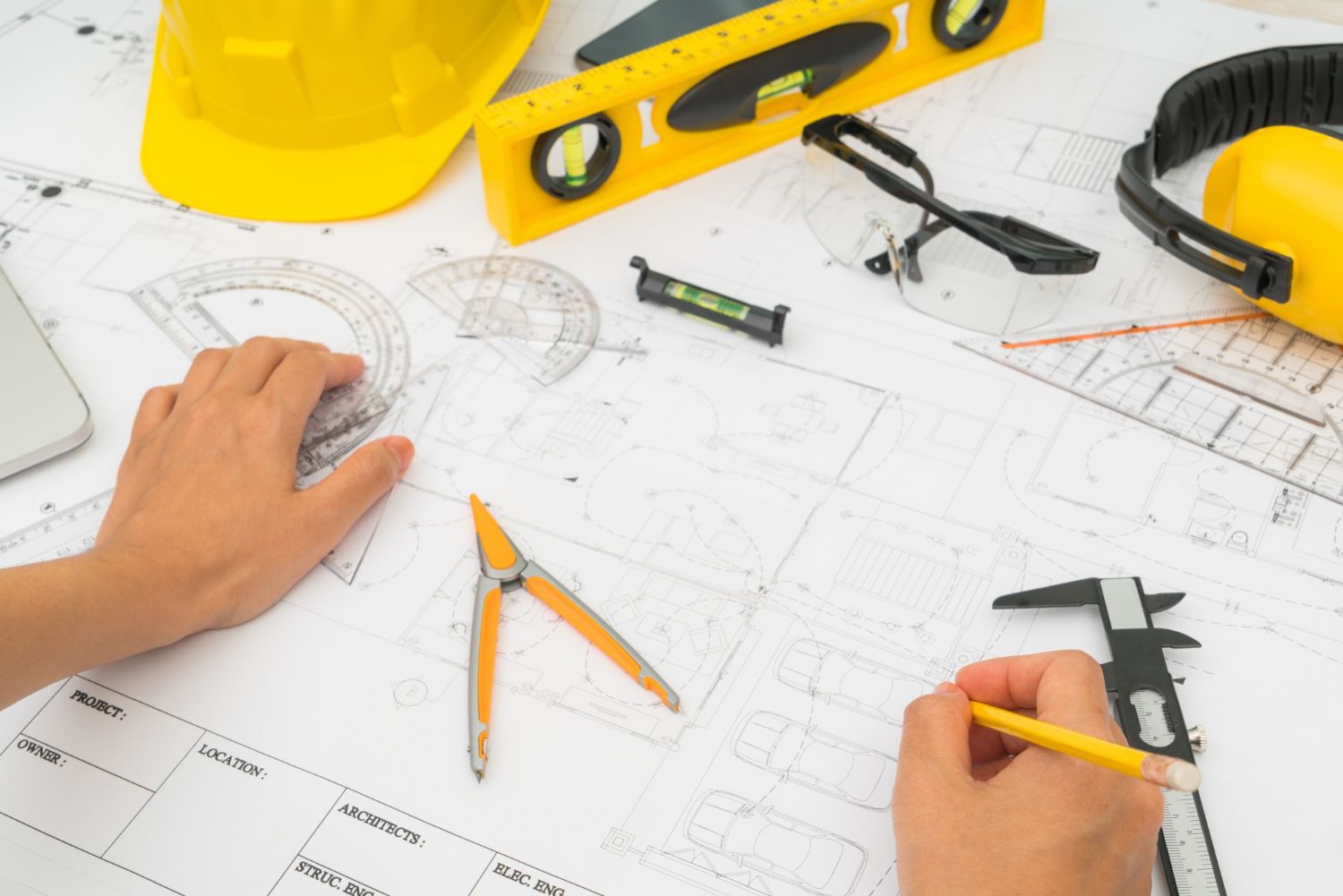 Manos utilizando herramientas sobre planos con equipo de seguridad, incluido casco y orejeras.