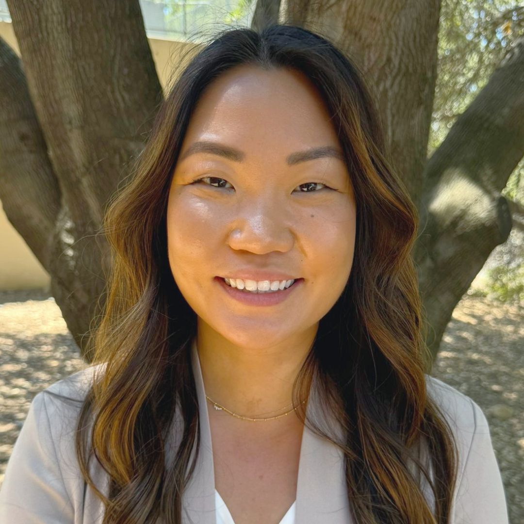 Woman with long dark hair smiles outdoors, wearing a light blazer and small gold necklace.