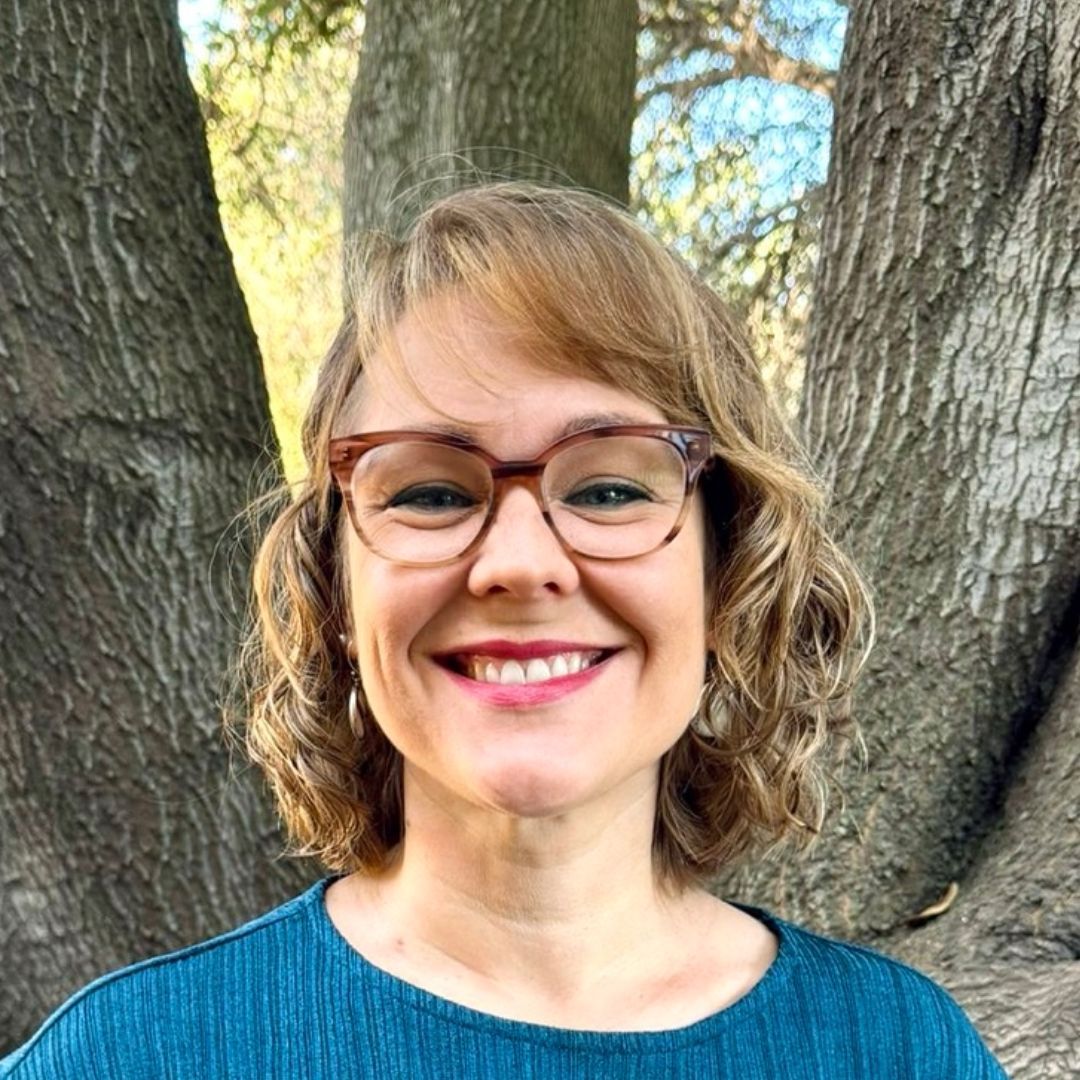 A woman wearing glasses and a blue shirt is smiling in front of a tree.