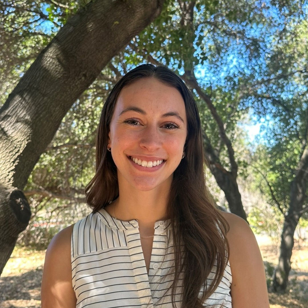 Woman with brown hair smiles, wearing a striped top, outdoors near trees on a sunny day.