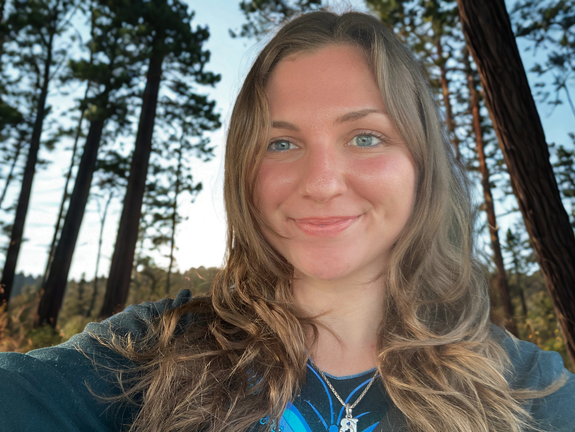 Woman smiles, looking at the camera. Trees in background.