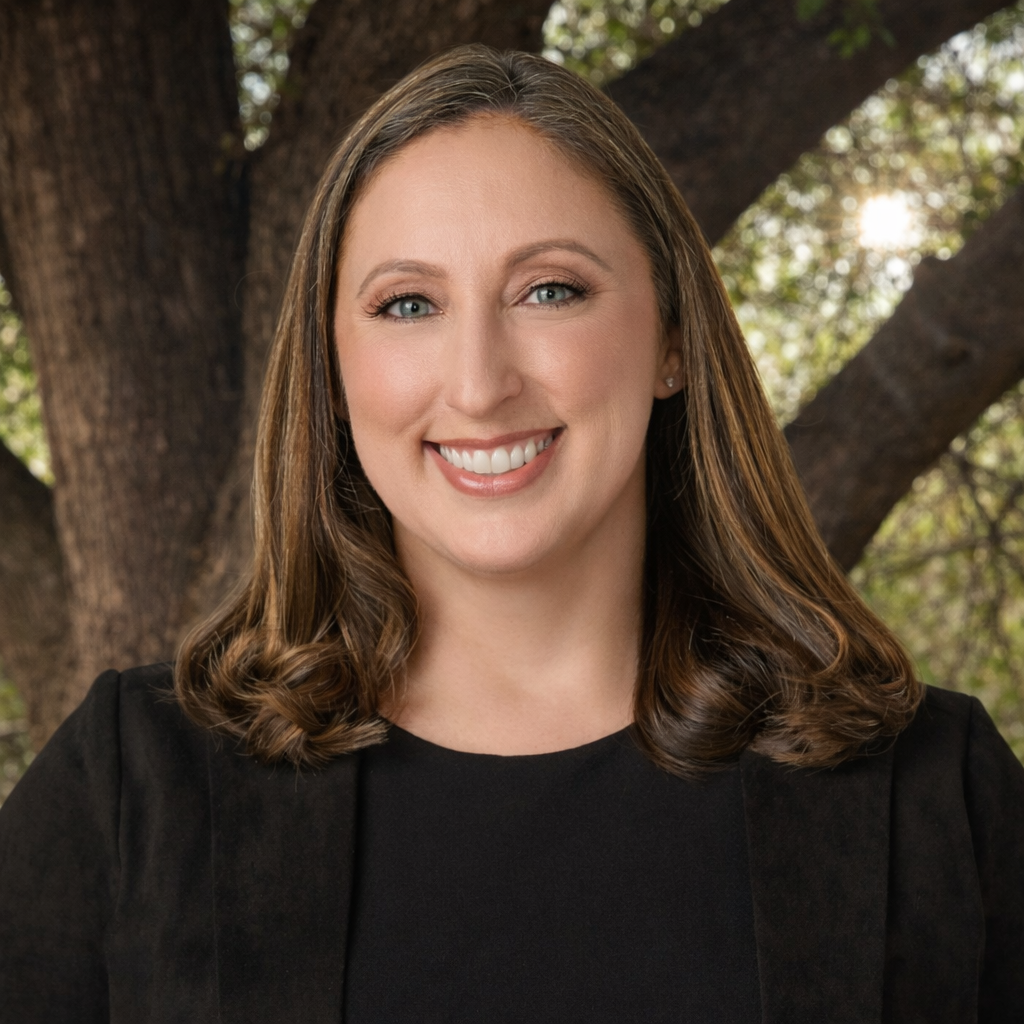 Woman with brown hair smiling, wearing a black blazer, standing outdoors.