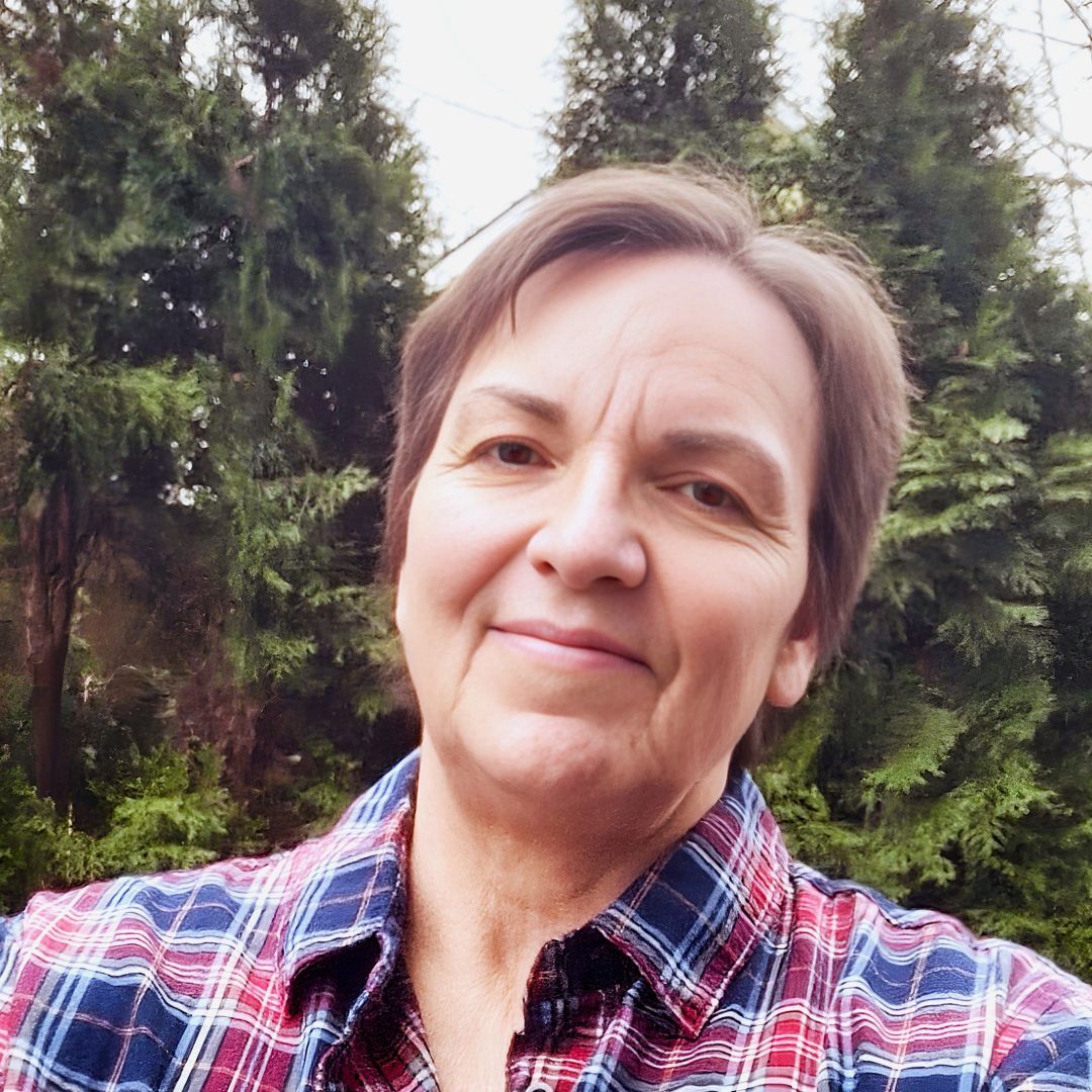 Woman with short brown hair smiles, wearing a plaid shirt. Trees are in the background.