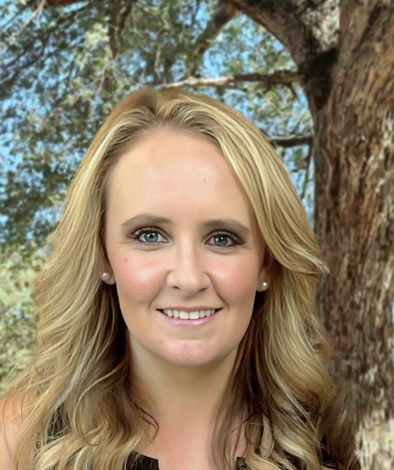 Blonde woman smiles at the camera, wearing pearl earrings, set against a tree-lined backdrop.