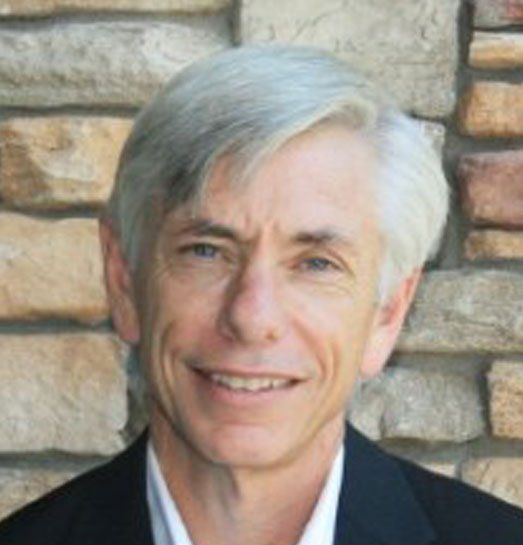 A man in a suit and white shirt is smiling in front of a stone wall.