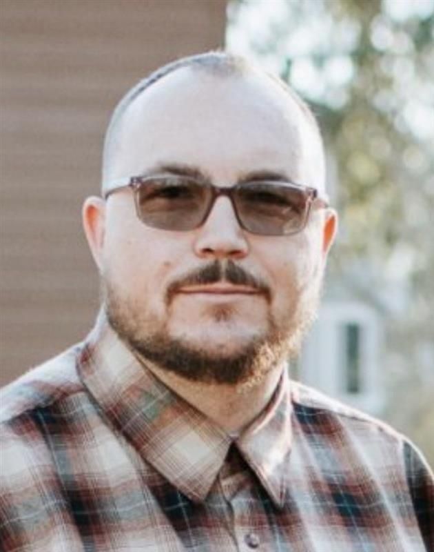 Man wearing glasses and plaid shirt, looking forward with a serious expression.