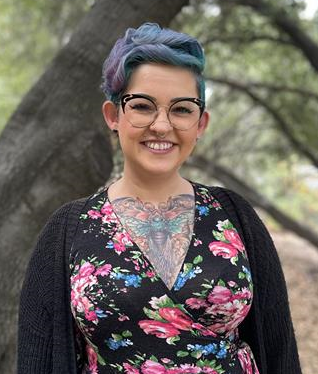 Woman with blue hair and glasses, smiling, wearing floral dress and black cardigan, outdoors near trees.