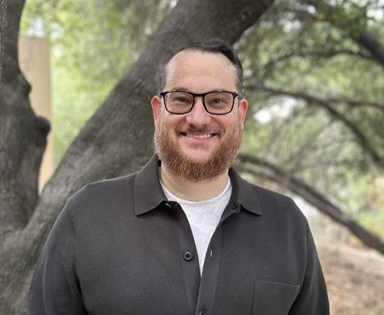 Man with beard and glasses smiling in front of a tree. He is wearing a dark button-up shirt over a white shirt.