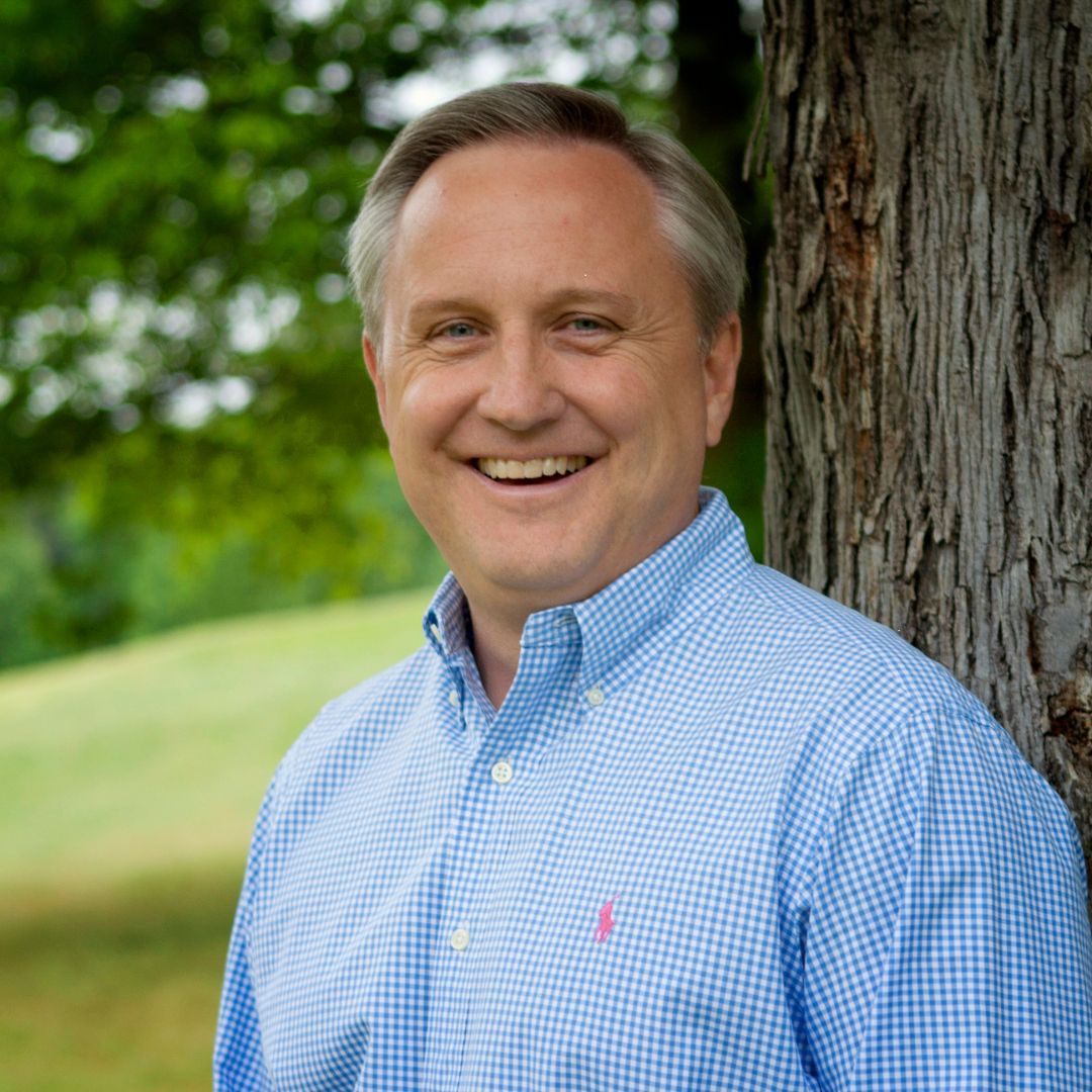 Man smiles, leans against tree, wearing blue checkered shirt with pink logo. Outdoors, daytime.