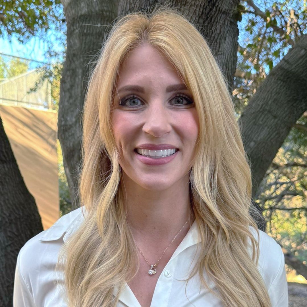 Blonde woman smiling, wearing a light-colored top, necklace. Outdoors with tree background.