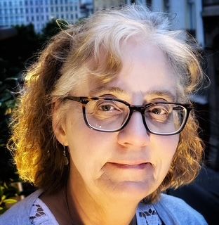 Woman wearing glasses, looking at the camera. She is lit from the front with a blurred background.