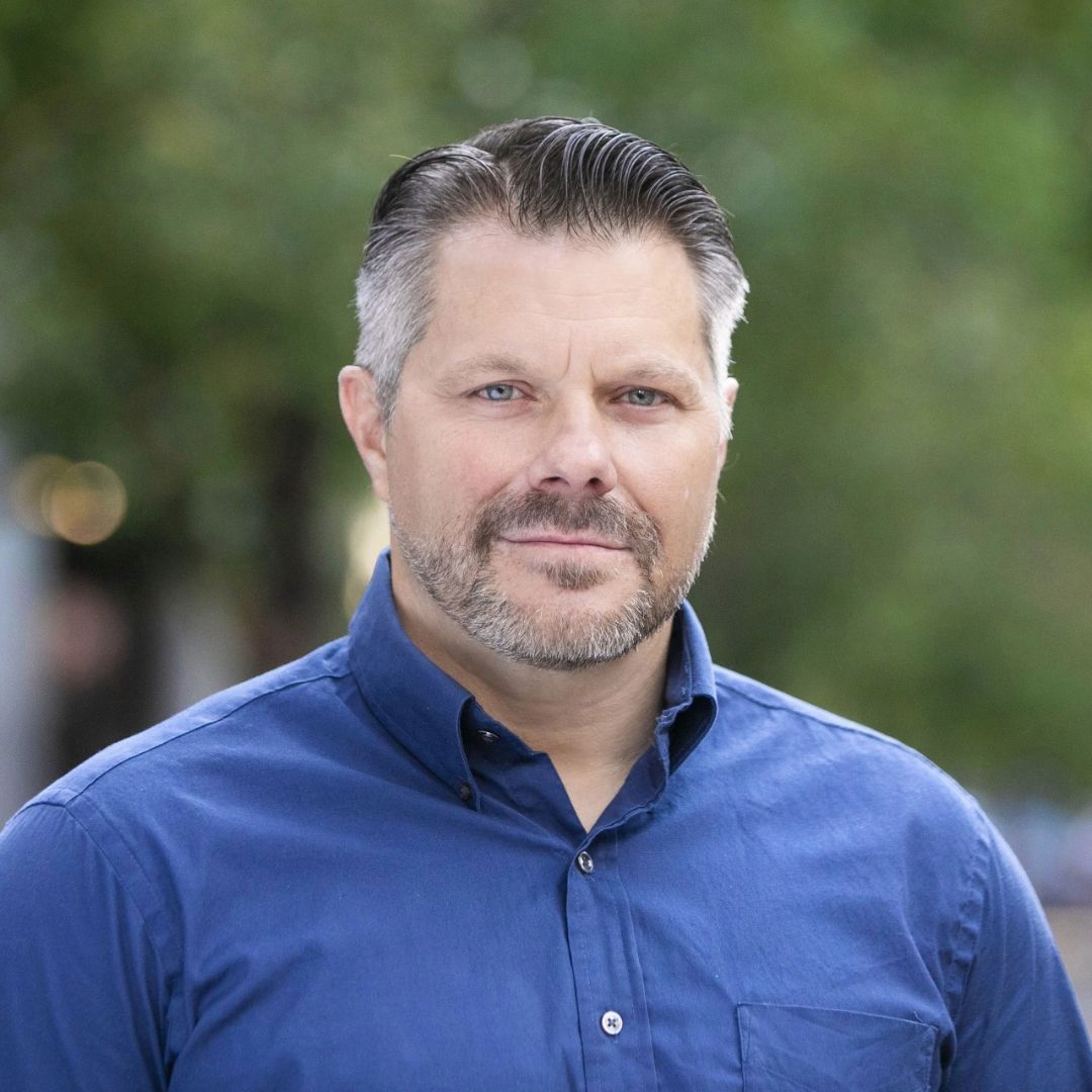 Man in blue shirt with a beard, outside, smiling.
