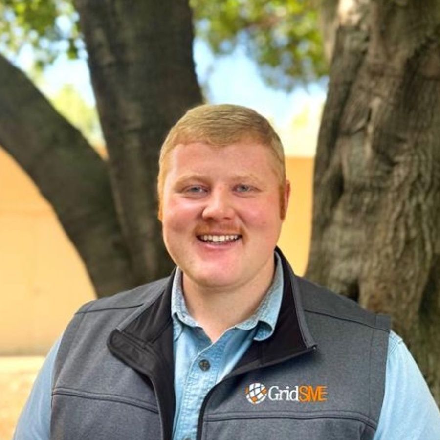 Man with blond hair and mustache wearing a gray vest with GridSME logo, smiling in front of a tree.