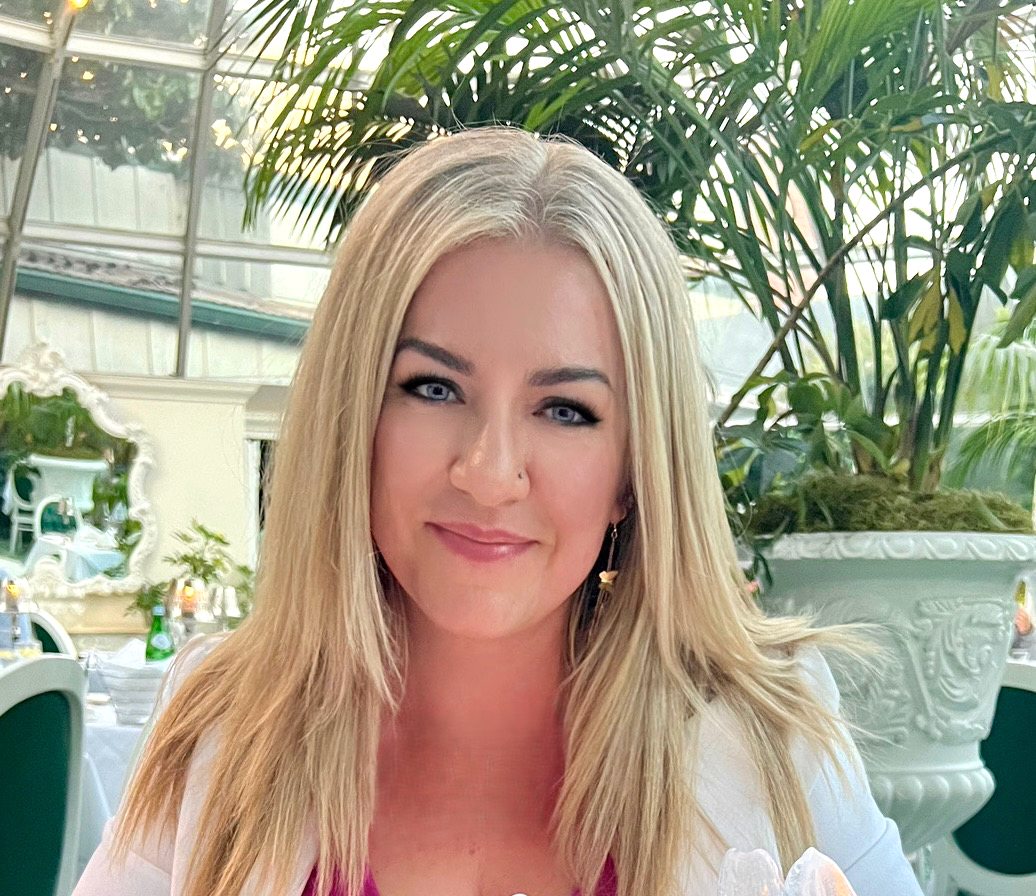 Woman with blonde hair smiles, seated at a table with a white jacket and tropical plants in the background.