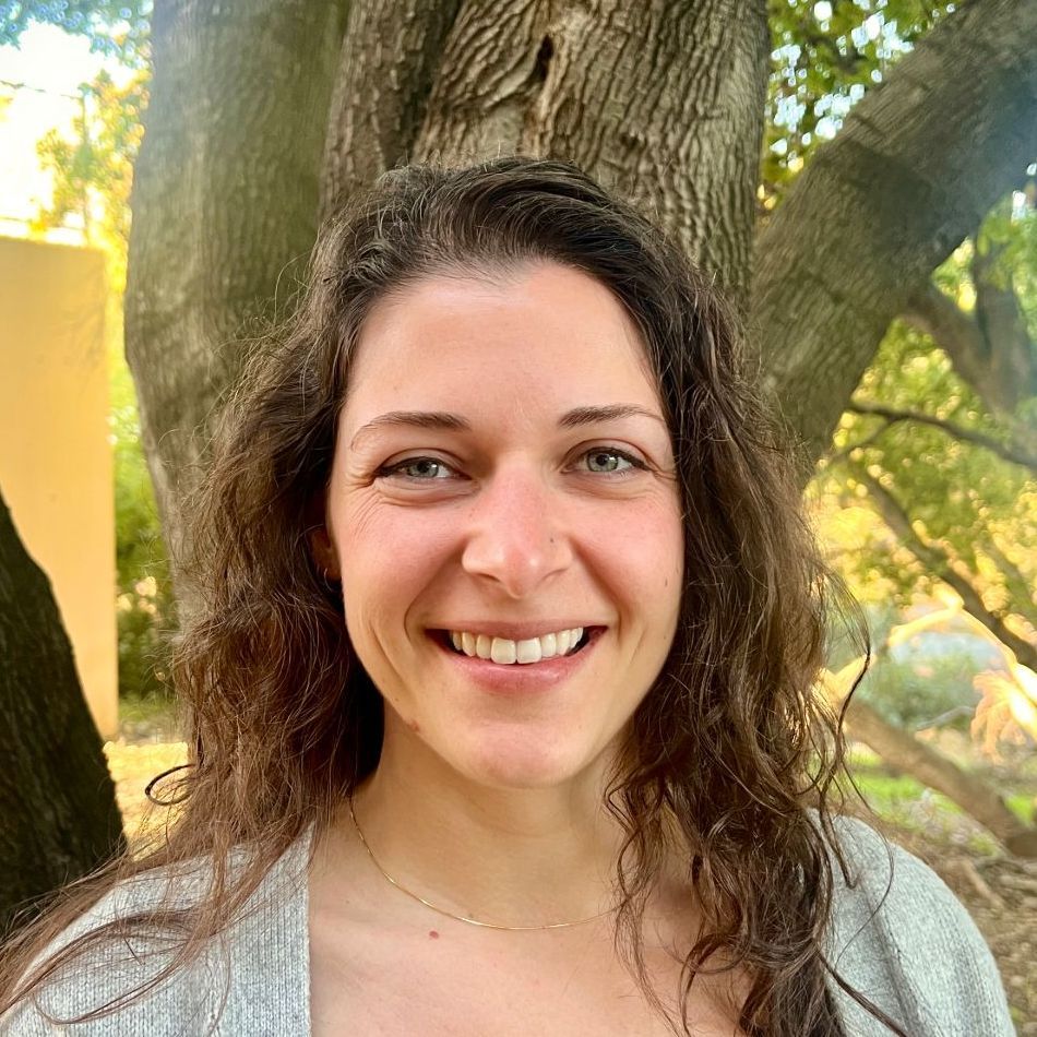 Woman with wavy brown hair smiles at the camera, in front of a tree, wearing a light gray cardigan.