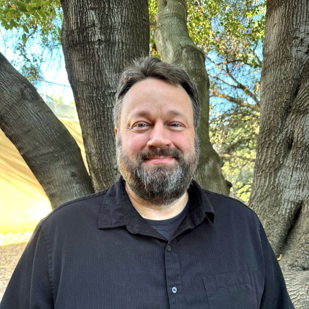 A man with a beard is standing in front of a tree.