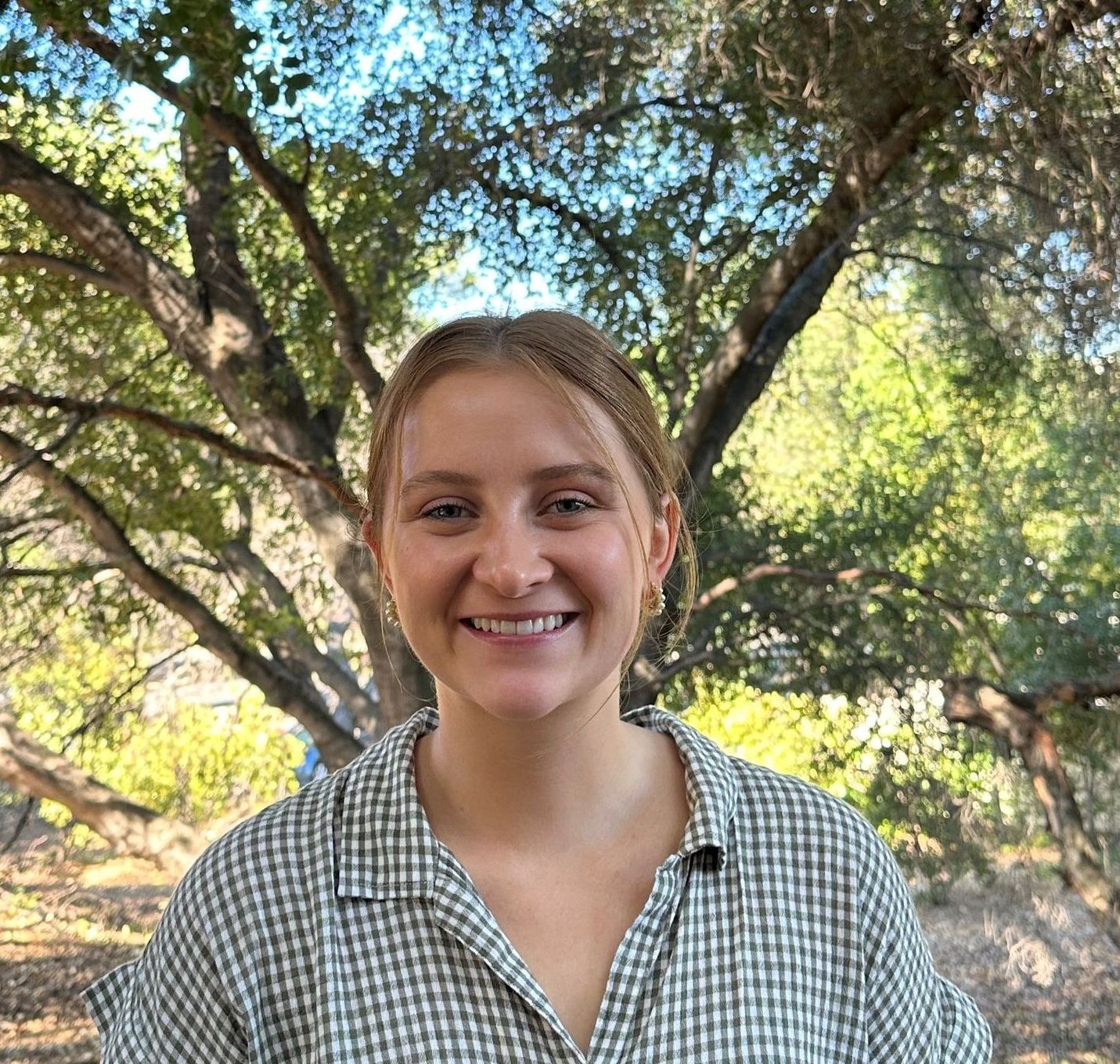 A woman in a plaid shirt is smiling in front of a tree.