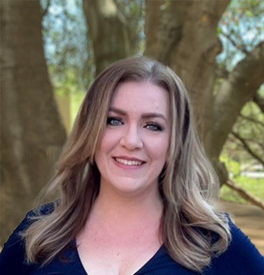 A woman in a blue shirt is smiling in front of a tree.