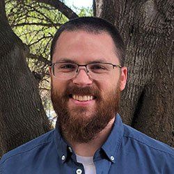 A man with a beard and glasses is smiling in front of a tree.
