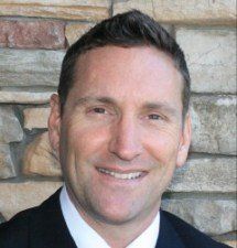 A man in a suit and tie is smiling in front of a stone wall.