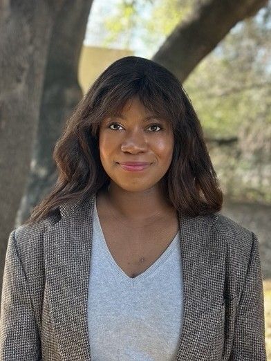 Woman in gray shirt and tweed blazer smiles in front of a tree.