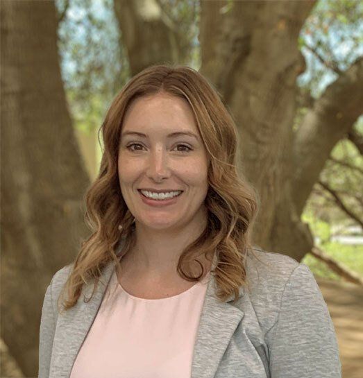 A woman in a pink shirt and gray jacket is smiling in front of a tree.