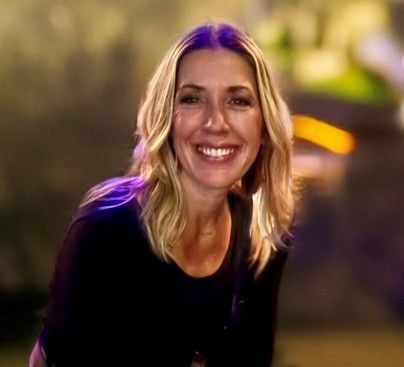 Smiling woman with blonde hair, wearing a black shirt, outdoors with blurred background and lighting.