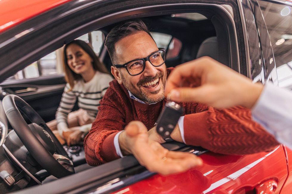 Un uomo sorridente riceve le chiavi da una mano in un'auto rossa, con una donna sullo sfondo.