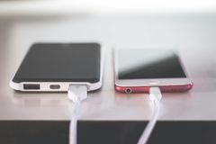 Zwei Smartphones, eines schwarz, eines rot, sind auf einer hellen Oberfläche mit weißen Ladekabeln verbunden.