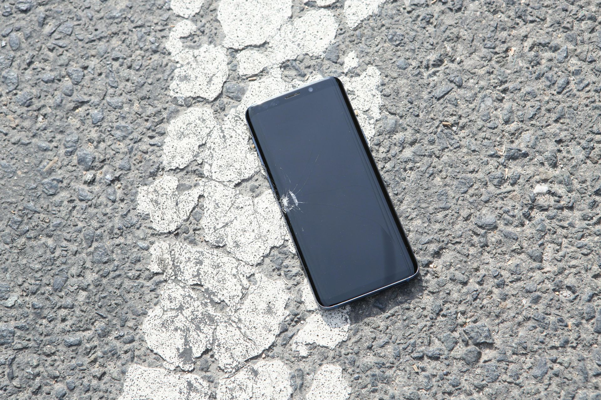 Schwarzes Smartphone liegt auf rissigem Asphalt mit einer weißen Linie.