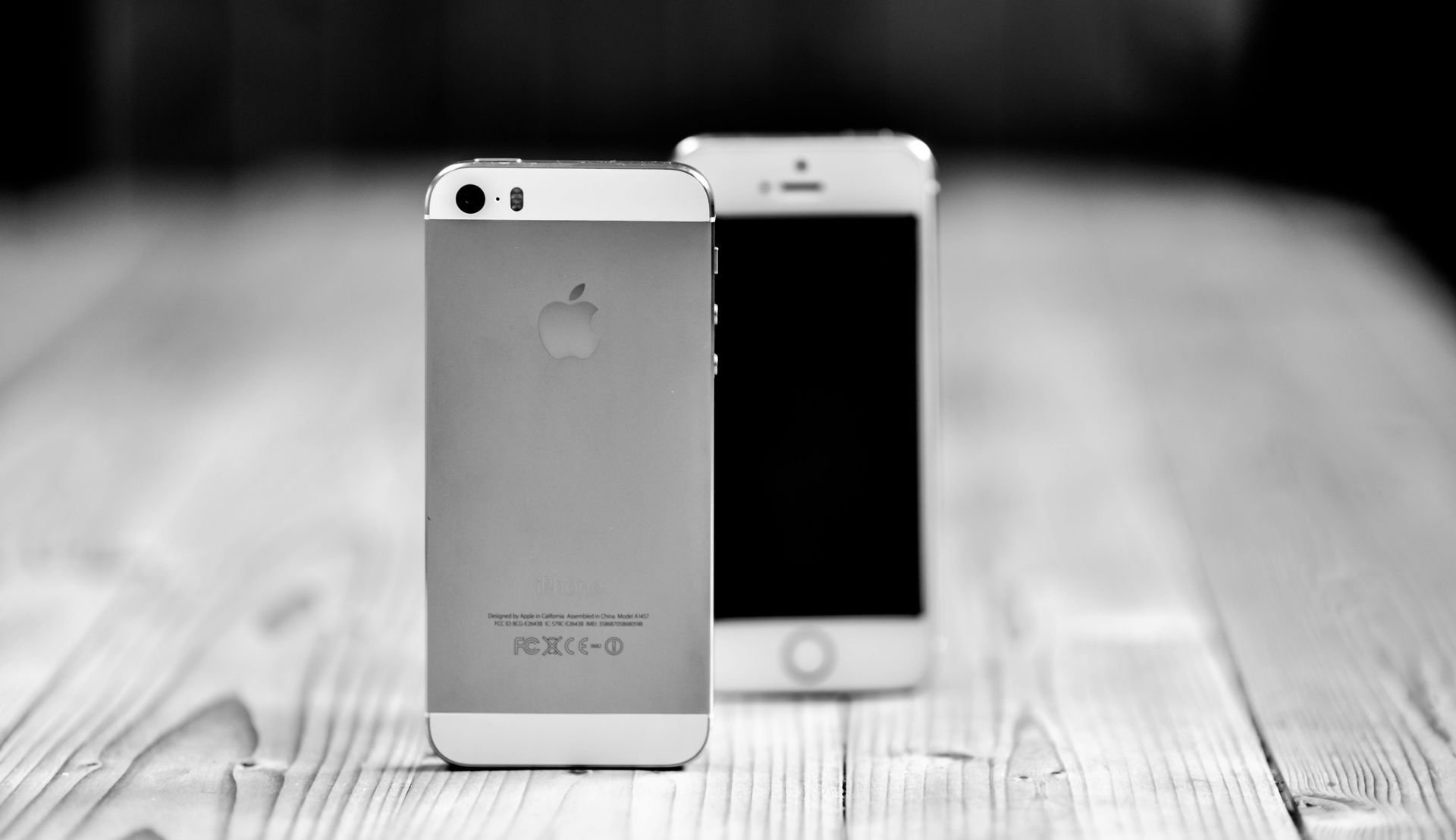 Zwei silber-weiße iPhones auf einer Holzoberfläche, eines mit ausgeschaltetem Bildschirm, das andere mit der Rückseite nach oben.