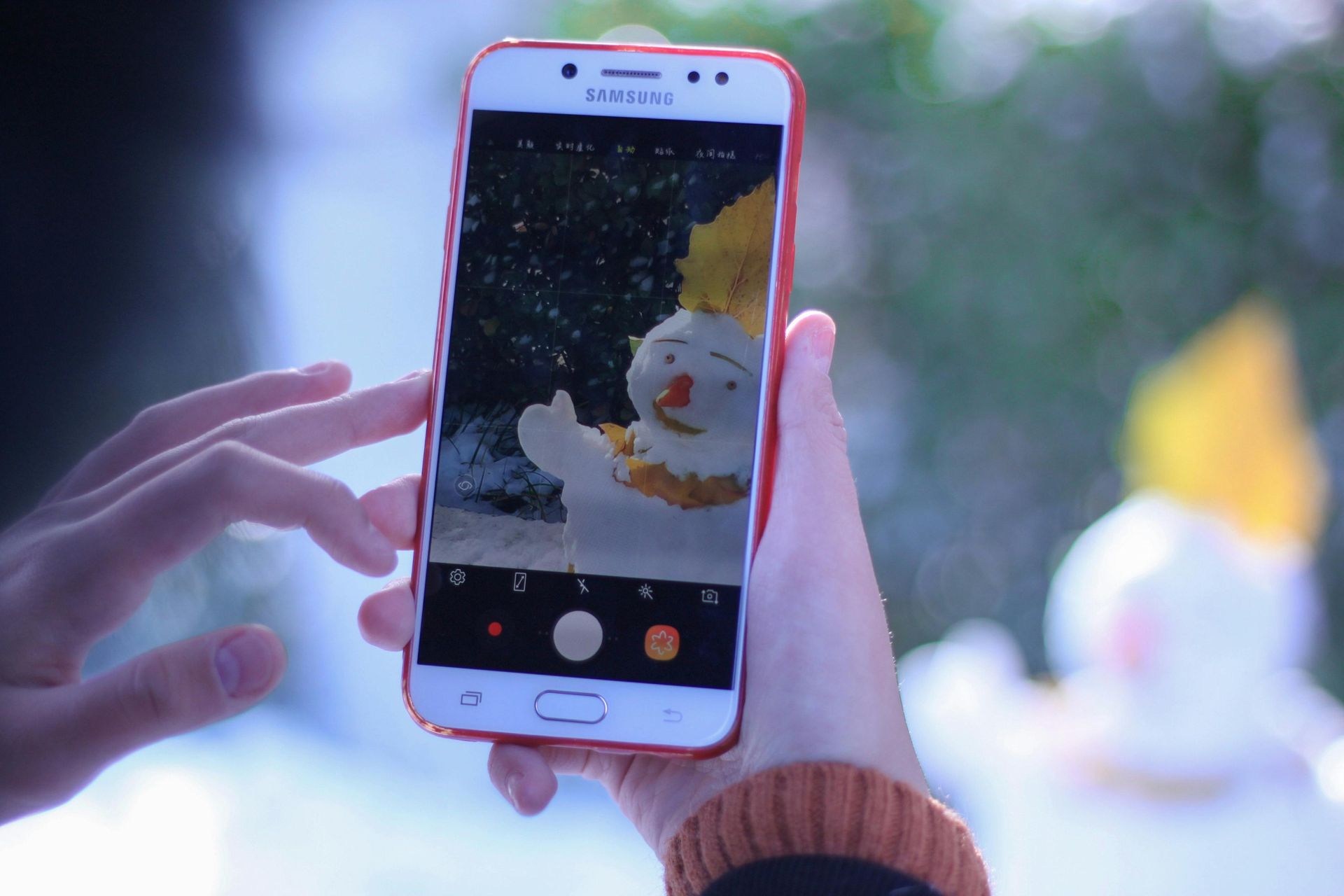 Eine Person hält ein Handy in der Hand und fotografiert eine Schneemann-Dekoration mit orangefarbenem Schal und Hut.
