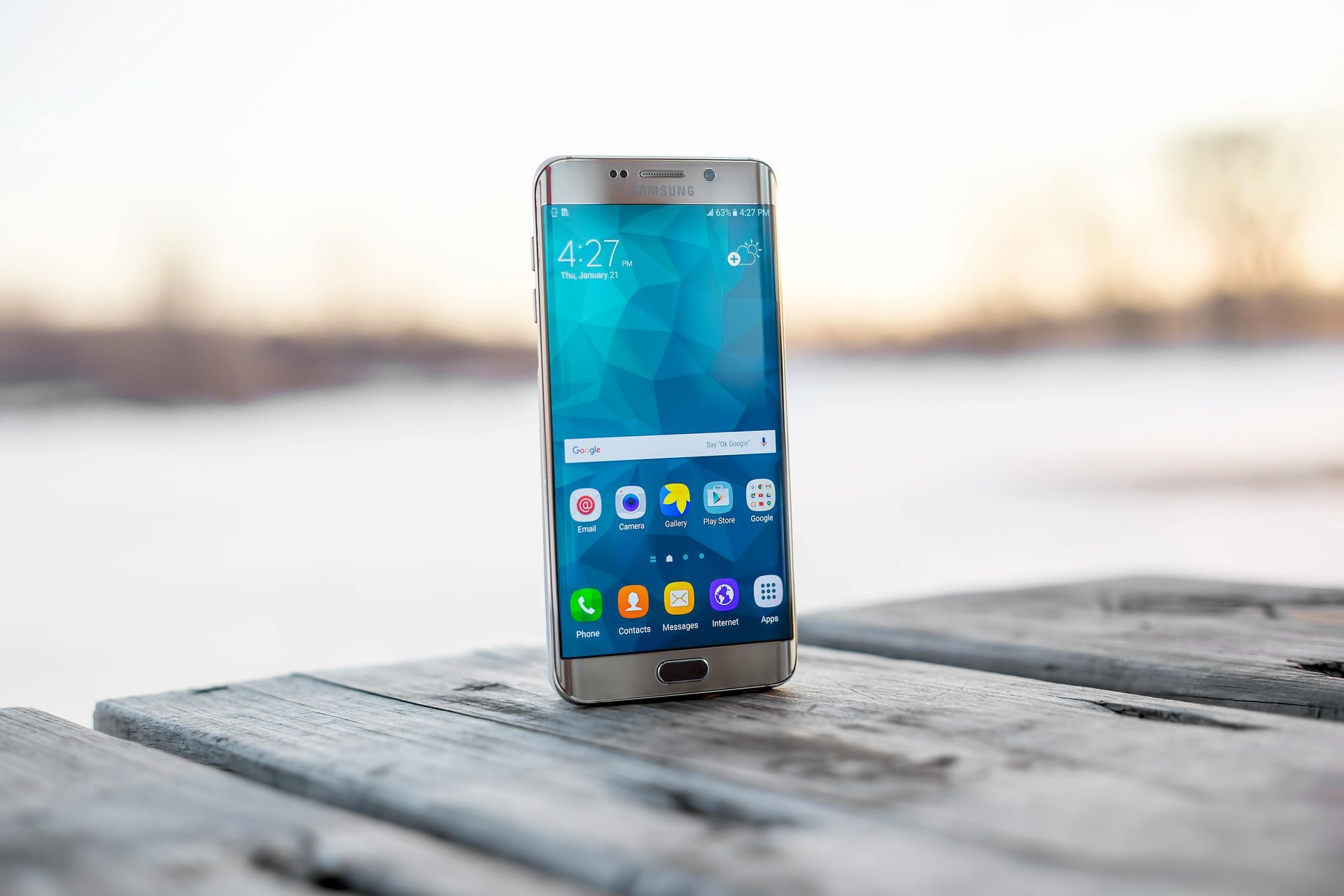 Silbernes Smartphone auf einer verwitterten Holzoberfläche, im Hintergrund eine verschwommene winterliche Außenkulisse.