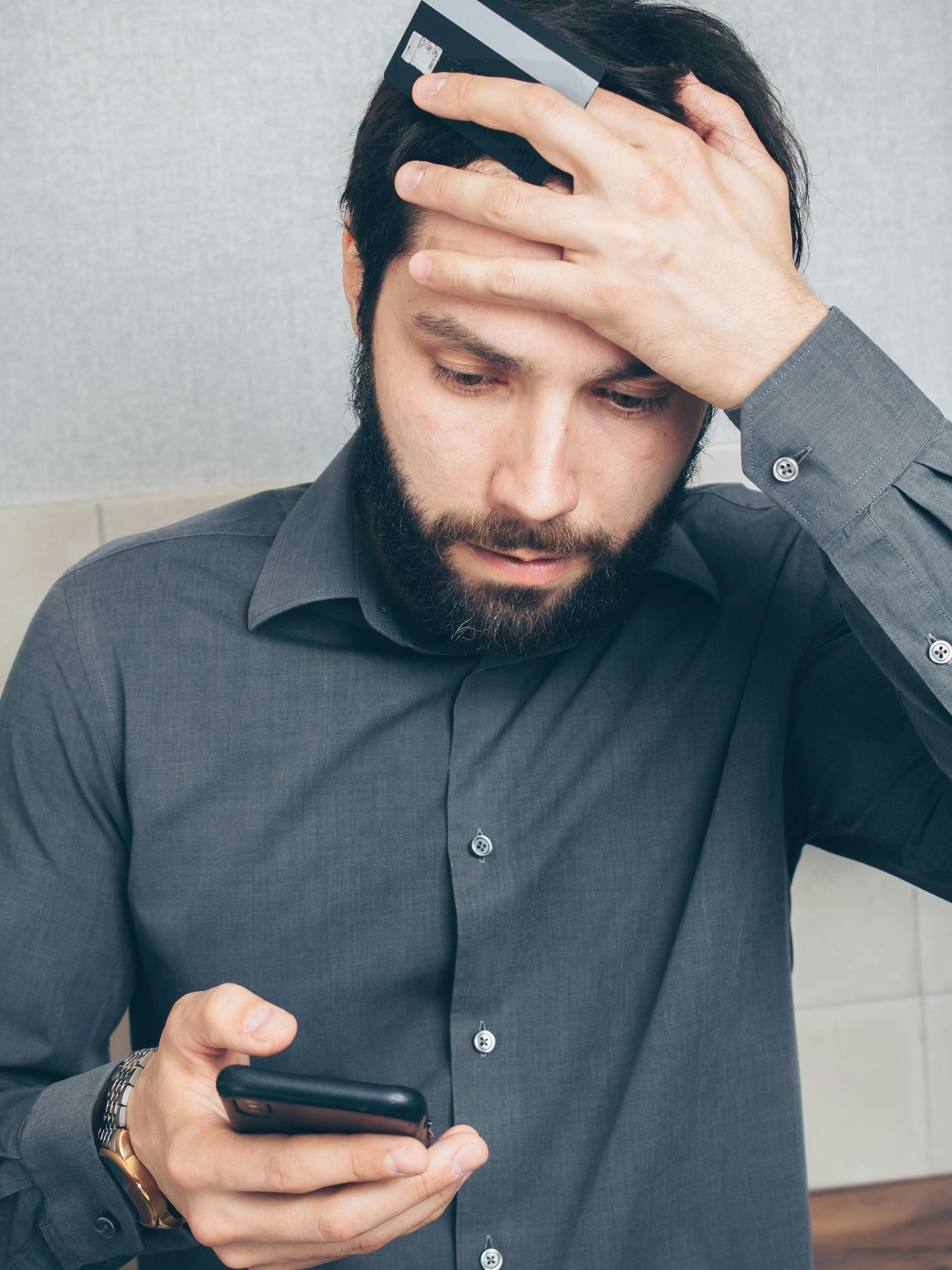 Ein Mann, der gestresst auf sein Handy starrt und sich eine Kreditkarte an den Kopf hält.